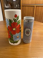 Two vases on wooden table; one tall white ceramic with large red and blue hand painted flowers, one shorter with fine floral pattern and flower medallion.