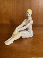 Front view of porcelain figurine depicting seated blonde girl with hands on knees, detailed facial features, painted clothing pattern, sitting on wooden surface.