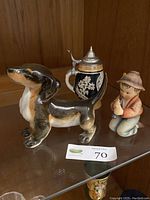 Full view of dachshund dog figurine, German beer stein, and Hummel Goebel Little Tooter figurine on a glass shelf.