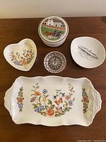Full lot shot of 5 vintage dishes on wood surface including floral porcelain pieces, covered porcelain dish, clear glass dish and ship motif dish.