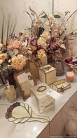 Overview photo showing multiple vases with faux flowers, decorative tissue holders, a hand mirror, and candles on a bathroom countertop