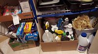 Cardboard box full of assorted cleaning supplies including spray bottles, bleach, and liquid containers. Boxes of household storage items like Ziploc bags and aluminum foil on shelf above.