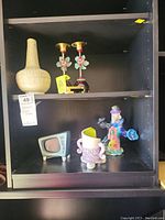 Shelf displaying six items: tall textured vase, two metal candle holders, ceramic TV model, ceramic clown figurine, and a small ceramic bowl-shaped item