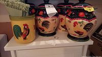 Photo showing the set of four ceramic canisters with rooster designs and a yellow ceramic container holding linens placed on a white shelf.