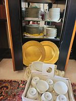 Image shows stacks of Noritake china plates and bowls inside a cabinet, two large gold serving platters in front, and a box of smaller bowls and cups.