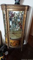 Full view of ornate gilt wood curio cabinet with glass panels and shelves, painted panels, and interior lighting