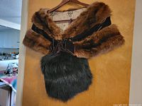 Black faux fur purse with brown velvet bow and strap, hanging on a wooden hanger against a tan background.