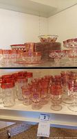 Glassware set on shelves showing colored top rims mostly pink to red color with clear glass bodies, various styles of glasses and bowls with some stacked plates.