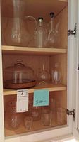 Glass cabinet with assorted clear glassware including oil pitchers, cheese dish with wooden base, and various drinking glasses and small containers.