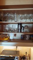 Three shelves filled with assorted stemware, glasses, and mugs including wine glasses, champagne coupes, small drinking glasses, several mugs, a glass jar, and a yellow container.
