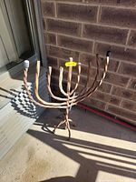 Iron menorah placed outside on a concrete surface next to a brick wall and door. Menorah shows twisted metal on its branches and base with visible rust and weathering.