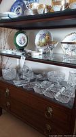 Front view of the tea cart showing three glass shelves loaded with crystal glassware and decorative plates on a wooden frame with brass handles on drawers below.