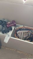 Photo showing a white box containing several hand weights of different sizes, a small woven basket with DVDs and other items.