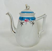 Front side profile of the white porcelain tea pot showing blue band with floral decorations and gold trim on spout, handle, and lid.