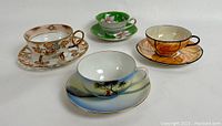 Set of four tea cups and saucers including green floral Trimont, Noritake winter landscape, orange citrus Art Deco Altrohlau, and footed gilded Japanese cup.