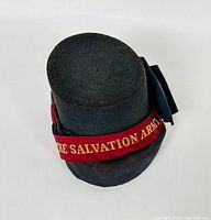 Top view showing woven black bonnet hat with red ribbon embroidered with 'THE SALVATION ARMY' in gold.