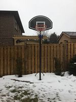 Full view of the basketball hoop stand around 12 feet tall in a backyard with snow on the ground and wooden fencing behind.