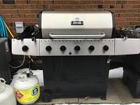 Front view of Broil-Mate propane BBQ grill with lid closed, showing six control knobs and two side shelves, outdoors near a brick wall.