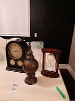 Overall view of four items including the Bombay sand clock, table clock, glass vase, and ceramic decor piece arranged on a white table