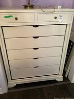 Front view of white painted wood cabinet showing the six drawers and brass lamp on top.