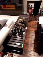 Top-down angled view of dark wooden chaise lounge with slatted top, two lion statues, and smaller items on it, placed on hardwood floor near a sofa and chairs.