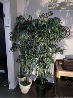 Photo of four tall faux plants with long, narrow green leaves and multiple green stems in plastic pots of different colors placed indoors near a wall.