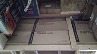 Eight cardboard boxes labeled 'In-Ceiling Sound Enclosure' by SpeakerCraft, stored in wooden rafters above garage area.