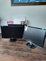 Two black computer monitors, one Samsung 24-inch and one ViewSonic 23-inch, pictured together on a wooden floor.