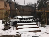 View of two stone planter pots with coniferous trees positioned near trampoline and wooden steps in snowy yard.