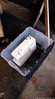 Janome JD1008 white sewing machine inside clear plastic bin, showing front with stitch dials and attached power cord