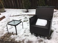 Outdoor dark brown metal woven chair with armrests and plastic covers on handles and top of backrest. Snow on chair seat where cushion is missing.