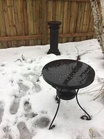 Photo showing black round metal outdoor table with perforated top and curved legs in snow with pedestal in background.