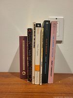 Books arranged standing in a row showing their spines against a wall with power outlet behind.