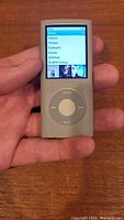 Front view of silver Apple iPod Nano 4th generation showing screen with menu options and click wheel, held in hand for scale.