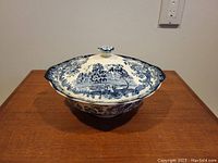 Front view of the covered vegetable dish showing the domed lid with scenic engraving and the flower knob handle on top.