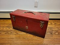 Closed view of old red metal toolbox showing latches, handle and surface with superficial rust.