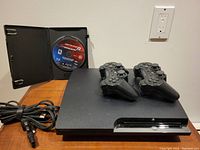Overall view of the Playstation 3 console with two controllers on top and game case to the left showing Need For Speed Hot Pursuit Limited Edition disc, and power cable coiled to the left.