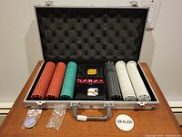 Open aluminum case containing poker chips in red, green, black, and white, two decks of cards, red dice, dealer button, and key, arranged neatly inside a padded foam liner.