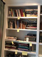 White bookshelf filled with assorted art and religious books, arranged on three shelves with some stacks and some upright, showing a variety of titles and colorful spines.