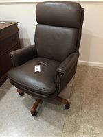 Full view of brown leather office chair with padded backrest, seat, armrests, and wooden base with caster wheels.