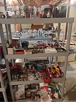 Wide view of multiple shelves containing assorted Christmas decorations including boxes, figurines, glassware, and ornaments in a basement setting.