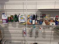 Shelf with multiple cleaning product bottles and boxes including bleach, Ajax spray, All detergent, WD-40, and various cleaning tools and bottles.