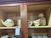 Wooden cabinet shelf containing Franciscan Rose tea set including stacked plates, teacups, and teapot with floral design