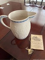 White Lenox ceramic pitcher with blue laurel leaf trim and embossed colonial/patriotic scenes, shown with original tag from Laszlo Ispanky.