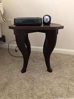 Wooden nightstand with a dark finish and curved legs, placed on a carpeted floor against a wall.