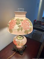 Side view of floral painted parlor lamp with glass shade and base, on ornate metal base with electrical cord.