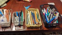 Photo showing four baskets of assorted pens, pencils, scissors, and tools on a table, displaying the variety of items in the lot.