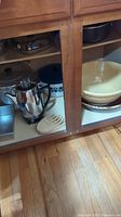Photo showing lower kitchen cabinet with stainless steel percolator, ceramic bowls, and glass-lidded pot inside wooden cabinet