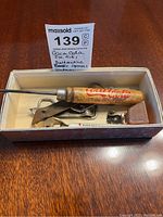 Box with vintage barware tools, including Coca-Cola branded wooden handle opener and other metal tools.