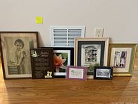 Seven framed artworks and photos arranged on a wooden surface leaning against a wall vent and power outlet, showing variety of sizes and styles.